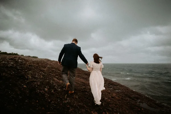 A bride and groom holding hands while walking along a rocky shoreline under a cloudy sky.