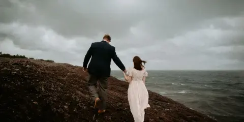 A bride and groom holding hands while walking along a rocky shoreline under a cloudy sky.