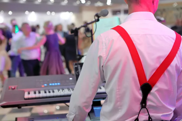 A musician in a white shirt and red suspenders plays a keyboard at a live event while guests dance in the background.