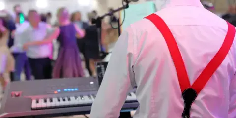 A musician in a white shirt and red suspenders plays a keyboard at a live event while guests dance in the background.