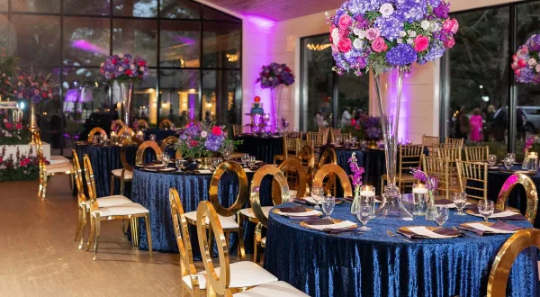 A wedding reception venue featuring round tables with blue velvet linens, gold chiavari chairs, and tall floral centerpieces.