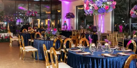 A wedding reception venue featuring round tables with blue velvet linens, gold chiavari chairs, and tall floral centerpieces.