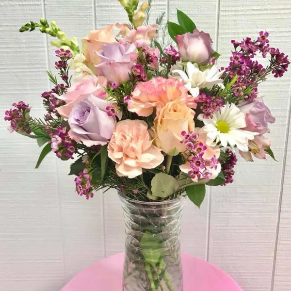A mixed bouquet of pastel roses, carnations, and purple filler flowers in a clear glass vase.