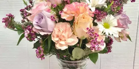 A mixed bouquet of pastel roses, carnations, and purple filler flowers in a clear glass vase.
