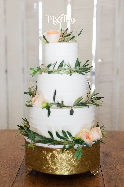A three-tiered white wedding cake decorated with greenery and peach roses, topped with a Mr & Mrs topper, sitting on a gold stand.