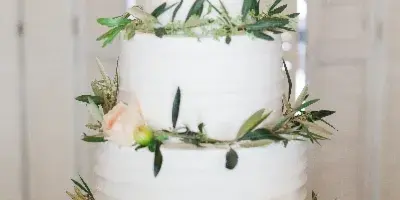A three-tiered white wedding cake decorated with greenery and peach roses, topped with a Mr & Mrs topper, sitting on a gold stand.