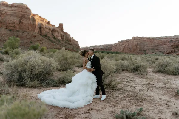 A bride in a white gown and a groom in a black suit sharing a kiss in a desert landscape with red rock formations.