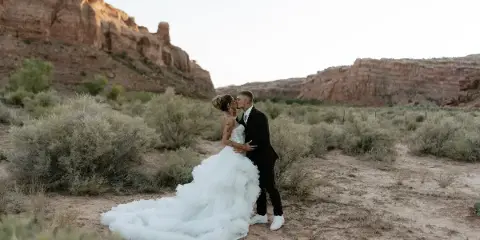A bride in a white gown and a groom in a black suit sharing a kiss in a desert landscape with red rock formations.