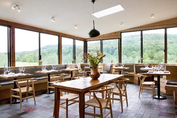 Interior view of a restaurant dining room with wooden tables, chairs, and large windows overlooking a forested valley.