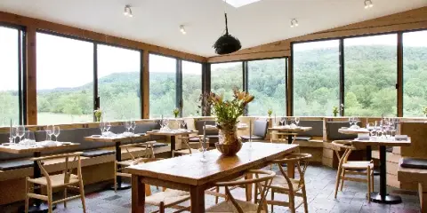Interior view of a restaurant dining room with wooden tables, chairs, and large windows overlooking a forested valley.