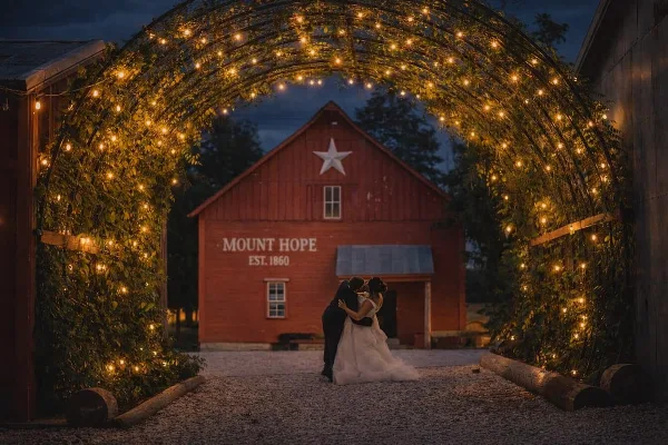 A bride and groom embracing in front of a red barn labeled 'MOUNT HOPE EST. 1860' under a string-lit archway at dusk.