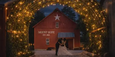 A bride and groom embracing in front of a red barn labeled 'MOUNT HOPE EST. 1860' under a string-lit archway at dusk.
