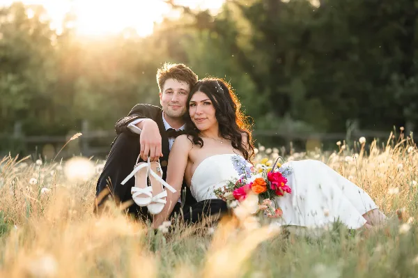 A bride and groom posing together in a grassy field at sunset, with the bride holding a bouquet of flowers.