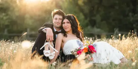 A bride and groom posing together in a grassy field at sunset, with the bride holding a bouquet of flowers.