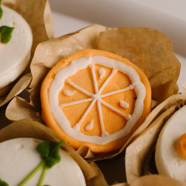 A close-up of a decorated orange cupcake with white icing piped in a citrus slice pattern, sitting in a brown paper liner.