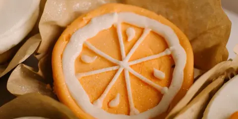 A close-up of a decorated orange cupcake with white icing piped in a citrus slice pattern, sitting in a brown paper liner.