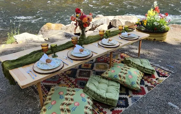 A vintage-style outdoor dining table set with patterned plates, glassware, and floral arrangements on a riverbank.