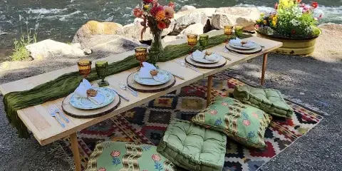 A vintage-style outdoor dining table set with patterned plates, glassware, and floral arrangements on a riverbank.