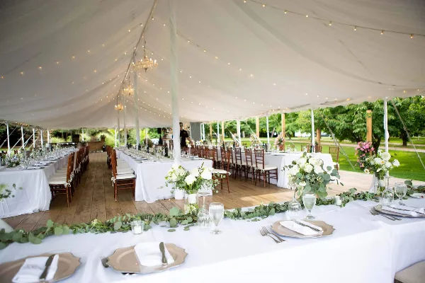 Interior view of a large white event tent set up for a wedding reception with long tables, chairs, and floral centerpieces.