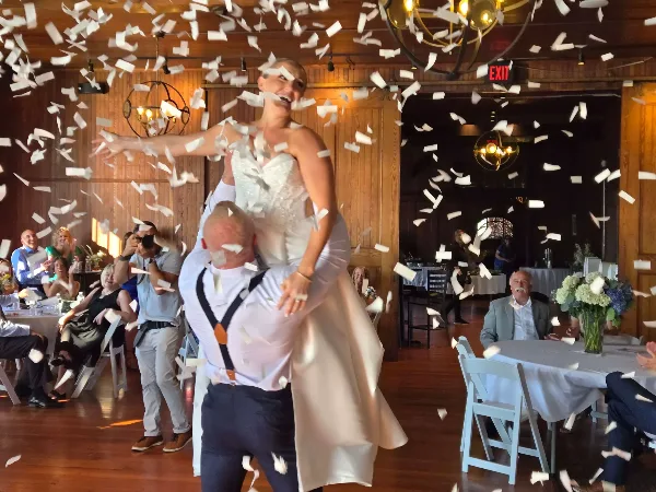 A groom lifts his bride onto his shoulders as they dance amidst falling confetti at a wedding reception.