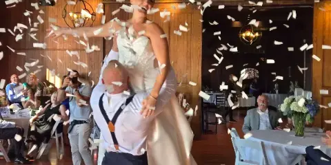 A groom lifts his bride onto his shoulders as they dance amidst falling confetti at a wedding reception.