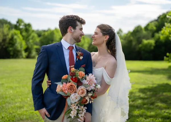 A bride and groom standing in a grassy field, looking at each other and smiling, with the bride holding a bouquet of flowers.