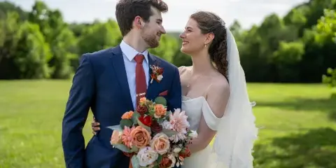 A bride and groom standing in a grassy field, looking at each other and smiling, with the bride holding a bouquet of flowers.