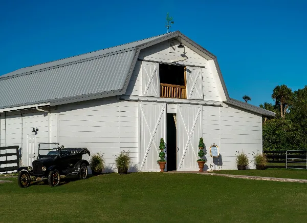 Exterior view of a white wooden barn with a metal roof, flanked by potted plants and an antique black car on a green lawn.