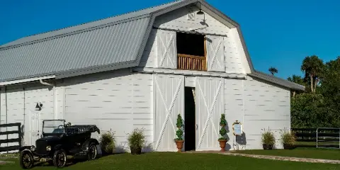 Exterior view of a white wooden barn with a metal roof, flanked by potted plants and an antique black car on a green lawn.