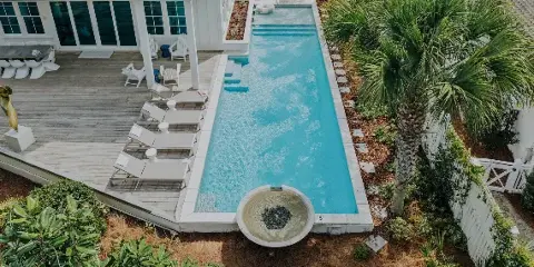 Aerial view of a luxury vacation home featuring a long swimming pool, wooden deck with lounge chairs, and palm trees.