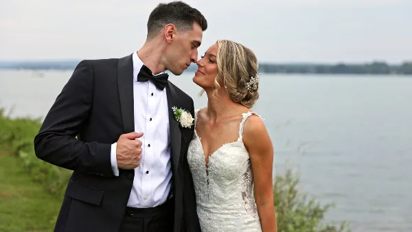Bride and groom in wedding attire standing close together outdoors with a lake in the background.