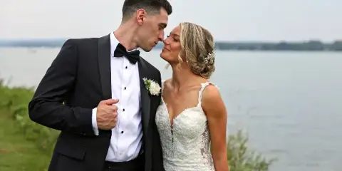 Bride and groom in wedding attire standing close together outdoors with a lake in the background.