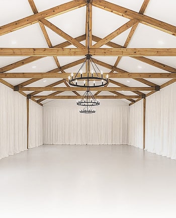 Interior view of a wedding venue with exposed wooden ceiling beams, white draping, and a tiered chandelier.