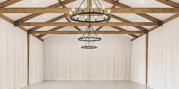 Interior view of a wedding venue with exposed wooden ceiling beams, white draping, and a tiered chandelier.