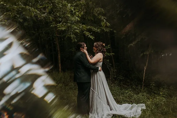 A wedding couple embracing in a forest setting, with the bride wearing a long grey dress and the groom in a dark suit.