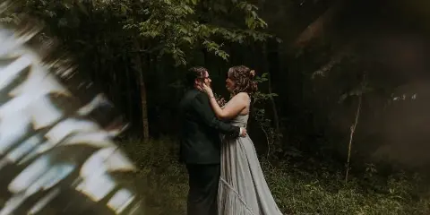 A wedding couple embracing in a forest setting, with the bride wearing a long grey dress and the groom in a dark suit.