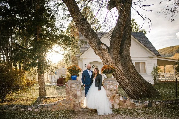 A bride and groom standing together in front of a white house with a large tree and stone pillars.