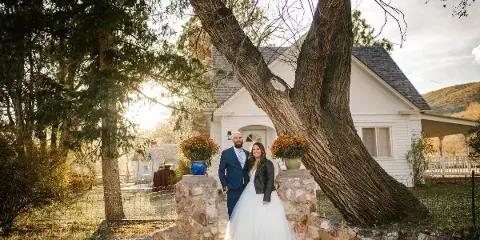 A bride and groom standing together in front of a white house with a large tree and stone pillars.