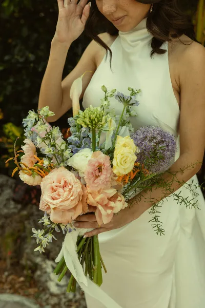 A bride in a white dress holding a pastel-colored floral bouquet with roses and greenery.