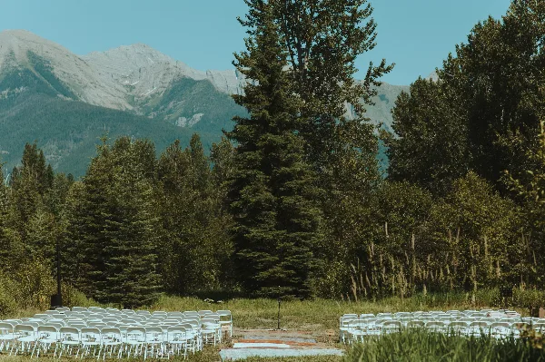 Outdoor wedding ceremony seating with white chairs in a grassy field, surrounded by evergreen trees and snow-capped mountains in the background.