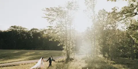 A bride and groom walking hand-in-hand through a sunlit grassy field with trees in the background.