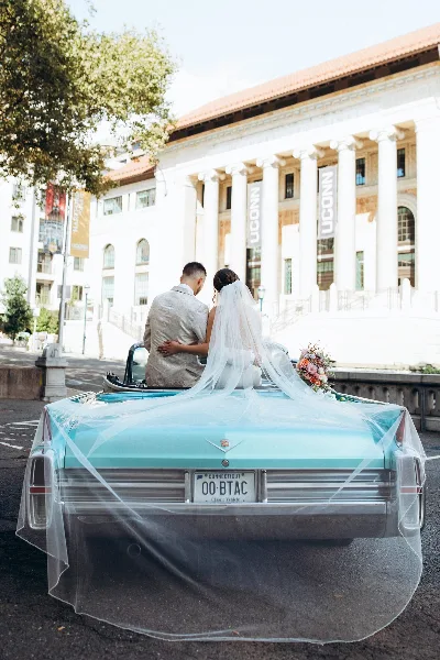 A bride and groom sitting in the back of a teal vintage convertible Cadillac with a veil and bouquet, parked in front of a building with columns.