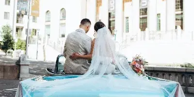 A bride and groom sitting in the back of a teal vintage convertible Cadillac with a veil and bouquet, parked in front of a building with columns.