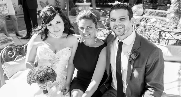 A wedding officiant sitting on a bench between a bride holding a bouquet and a groom in a suit.
