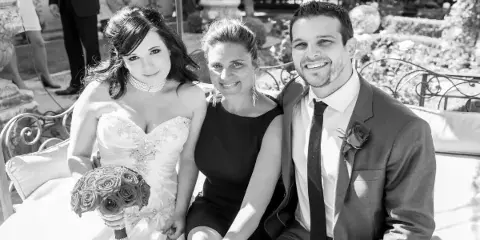 A wedding officiant sitting on a bench between a bride holding a bouquet and a groom in a suit.