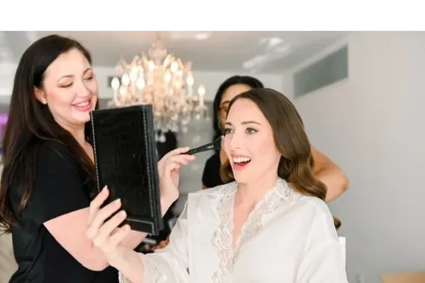 A makeup artist applying makeup to a smiling client in a white robe.