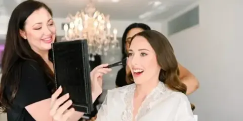 A makeup artist applying makeup to a smiling client in a white robe.