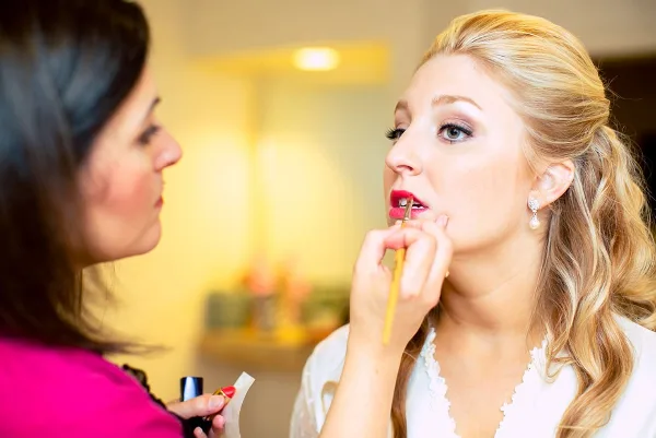 A makeup artist applies red lipstick to a blonde client with styled hair and pearl earrings.
