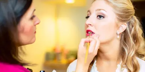A makeup artist applies red lipstick to a blonde client with styled hair and pearl earrings.