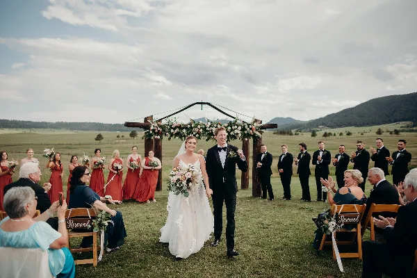 A bride and groom walking down the aisle at an outdoor wedding ceremony in a grassy field with mountains in the background.